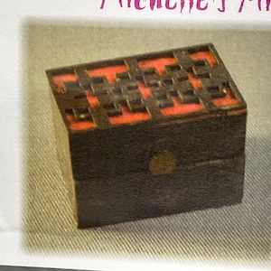 May include: A small, rectangular wooden box with a dark brown exterior and a red interior. The lid features a decorative, openwork design. A small, gold-colored circular clasp is on the front of the box. The box appears to be a miniature or a jewelry box.