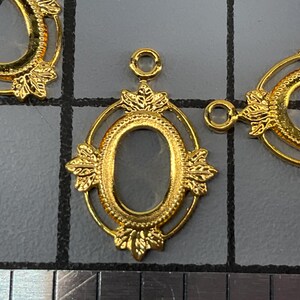 Op de afbeelding: Drie goudkleurige ovale metalen hangerframes met decoratieve bladachtige accenten aan de boven- en onderkant. Elk frame heeft een kleine ronde lus aan de bovenkant voor bevestiging. Een liniaal is onderaan zichtbaar met een schaalverdeling in cm.