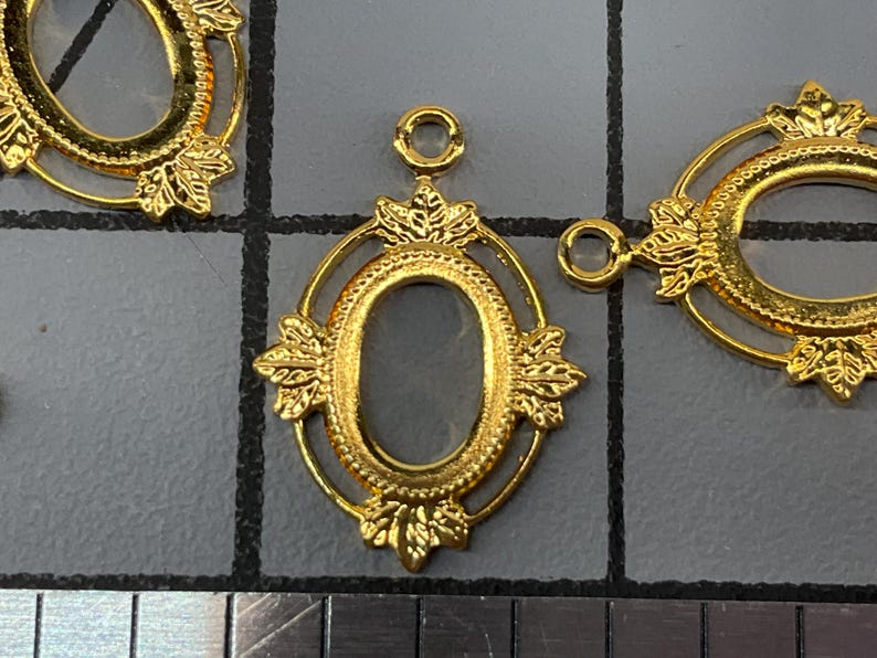 Op de afbeelding: Drie goudkleurige ovale metalen hangerframes met decoratieve bladachtige accenten aan de boven- en onderkant. Elk frame heeft een kleine ronde lus aan de bovenkant voor bevestiging. Een liniaal is onderaan zichtbaar met een schaalverdeling in cm.