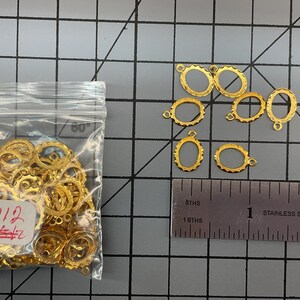 Op de afbeelding: Een verzameling kleine goudkleurige ovale metalen frames met geschulpte randen, elk voorzien van een klein lusje aan de bovenkant. Een doorzichtig plastic zakje bevat vele van deze frames en een liniaal toont een breedte van ongeveer 1,3 cm.