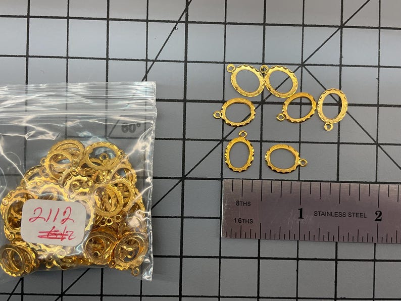 Op de afbeelding: Een verzameling kleine goudkleurige ovale metalen frames met geschulpte randen, elk voorzien van een klein lusje aan de bovenkant. Een doorzichtig plastic zakje bevat vele van deze frames en een liniaal toont een breedte van ongeveer 1,3 cm.
