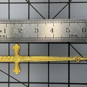 Może przedstawiać: Złoty metalowy wisiorek w kształcie miecza ze skomplikowanymi teksturowanymi detalami na rękojeści, jelcu i ostrzu. Miecz znajduje się pod metalową linijką, wskazującą całkowitą długość około 6,5 centymetra od czubka do głowicy.