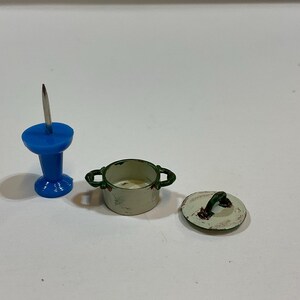 Puede incluir: Un chinche azul, una olla blanca en miniatura con asas verdes y una tapa a juego con un asa verde están dispuestas sobre una superficie blanca. La olla y la tapa parecen ser parte de un juego de casa de muñecas.