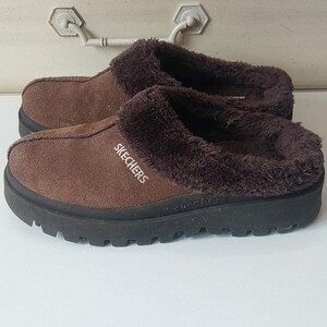 May include: Brown suede slip-on clogs with a plush, dark brown faux fur lining. The clogs have a black rubber sole and the brand name "SKECHERS" is printed in white on the side.