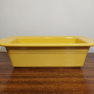 May include: A rectangular yellow ceramic baking dish with a smooth, glossy finish. The dish features a wide, flat rim and a slightly tapered base with a subtle horizontal ridge detail along the upper exterior side walls of the vessel.