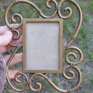 May include: Ornate, antique-style photo frame in a bronze finish. The frame features a rectangular opening for a photo, surrounded by decorative scrollwork and embellishments. The frame is held by a hand, with a blurred green background.