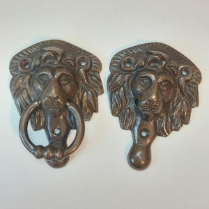 May include: Two antique bronze lion head door knockers. One knocker has a ring for knocking, the other is a matching lion head. The lion heads have detailed manes and are mounted on a rectangular backplate.