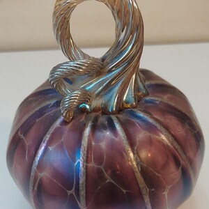 May include: A decorative glass pumpkin with a silver-colored, twisted handle. The pumpkin is a deep purple color with iridescent blue and silver accents. The pumpkin's surface has a textured, web-like pattern.