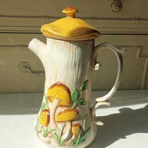 May include: A ceramic coffee pot with a cream-coloured tree trunk design, featuring yellow and brown mushrooms and green leaves. The lid is yellow, and the handle is curved. The pot is approximately 25 cm tall.