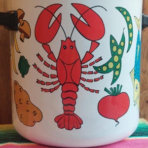 May include: White enamel cooking pot with a red lobster and various vegetables design. The pot features black handles and a silver rim. The design includes a red lobster, green peas, a red tomato, and other vegetables.