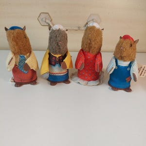 May include: Four small anthropomorphic animal figurines with fuzzy brown heads and fabric clothing. The figures wear various outfits including a blue cap, a white bonnet, a red dress, and blue overalls. Each figurine has distinct facial features.
