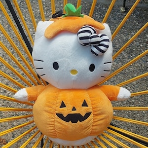 May include: A plush Hello Kitty doll dressed as a pumpkin for Halloween. The doll has a white head with black eyes and whiskers, a striped bow, and a pumpkin stem. The orange pumpkin body has a carved jack-o'-lantern face.