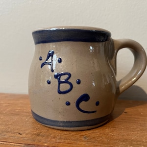 May include: A beige ceramic mug with a blue rim and handle. The mug has blue lettering that says "A.B.C." with dots around each letter.
