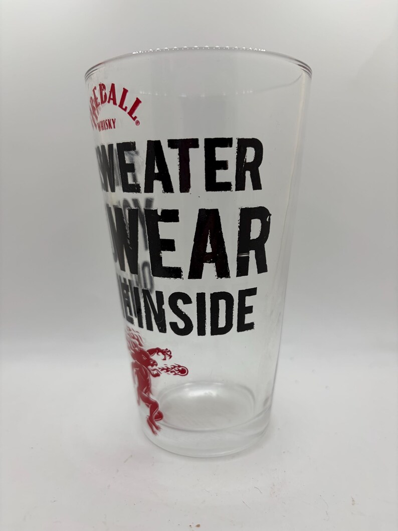 Puede incluir: Vaso de pinta transparente con el texto negro "WEATER WEAR THE INSIDE". El logotipo de Fireball Whisky est&aacute; en la parte superior y un gr&aacute;fico de drag&oacute;n rojo en la parte inferior. El vaso est&aacute; vac&iacute;o.