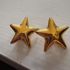 May include: Two gold-colored star-shaped earrings. Each earring has five points and a shiny, reflective surface. The earrings are displayed on a white surface.