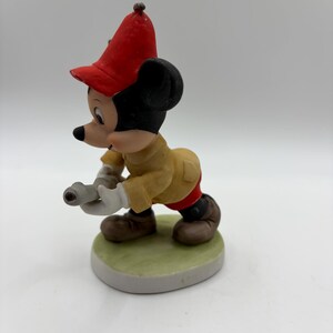 May include: A ceramic figurine of Mickey Mouse in a firefighter's uniform. Mickey is wearing a red hat, tan jacket, red pants, and brown shoes. He is holding a hose and standing on a green and white base.