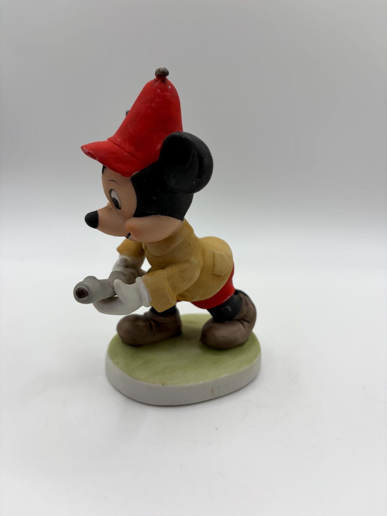 May include: A ceramic figurine of Mickey Mouse in a firefighter's uniform. Mickey is wearing a red hat, tan jacket, red pants, and brown shoes. He is holding a hose and standing on a green and white base.