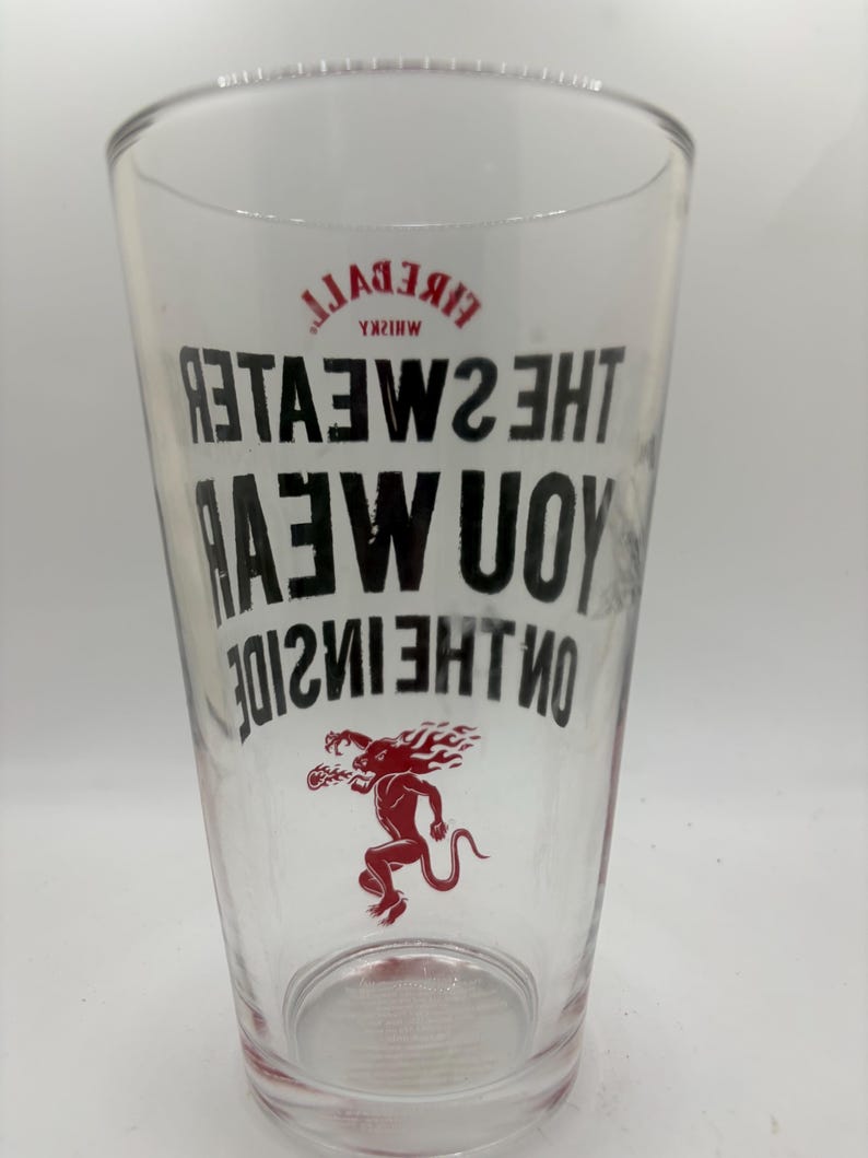 Puede incluir: Un vaso de pinta transparente con las palabras "Fireball Whisky" y "The Sweater You Wear On The Inside" impresas en negro. Tambi&eacute;n hay una ilustraci&oacute;n roja de una criatura m&iacute;tica.