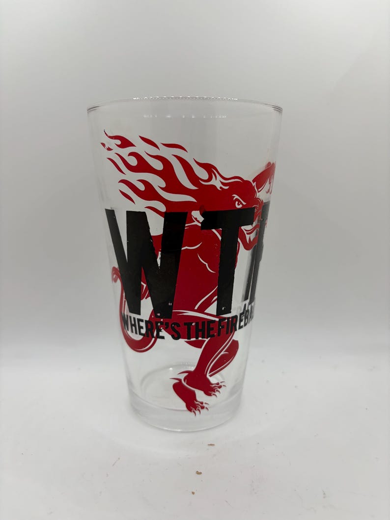May include: Clear glass pint glass with a red dragon and flame design. The glass features the black text "WTF" and "WHERE'S THE FIRE?". The glass is cylindrical and suitable for beverages.
