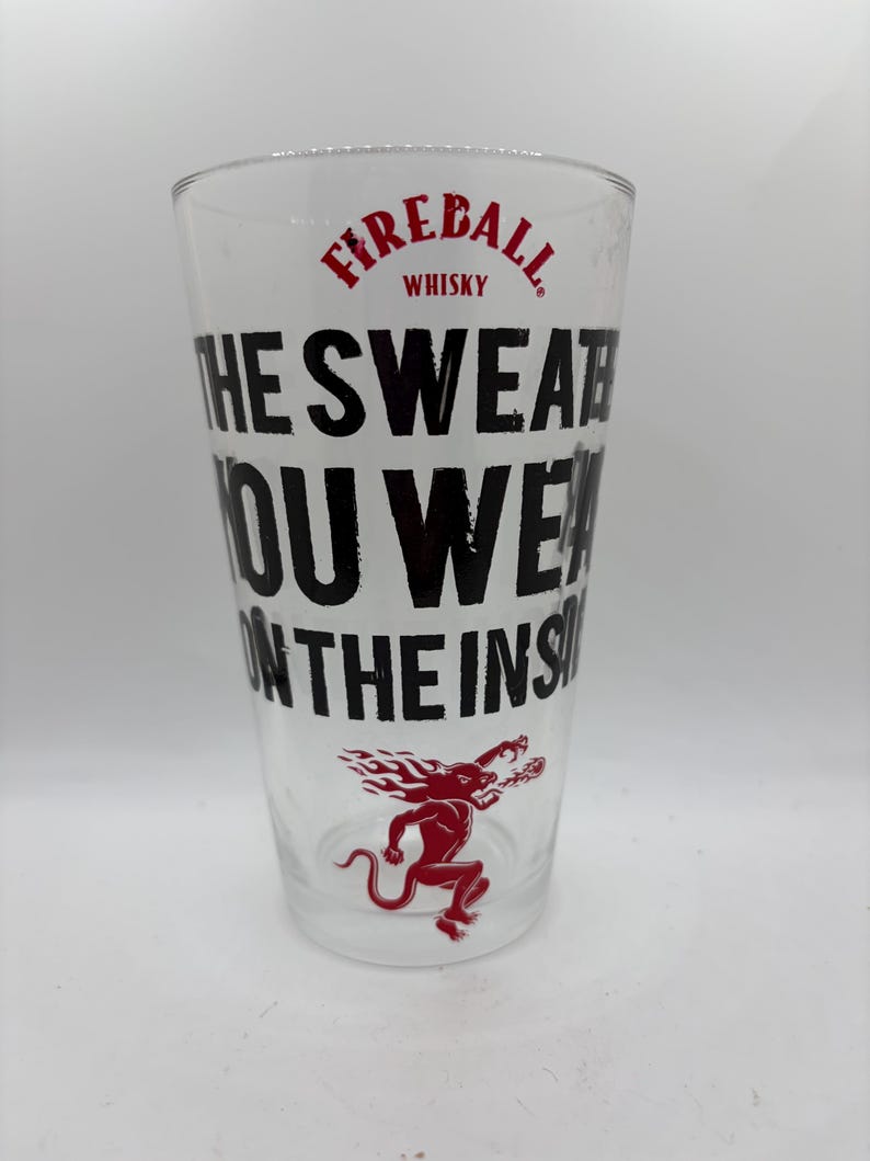 Puede incluir: Un vaso de pinta transparente con el texto "THE SWEAT YOU WEAR ON THE INSIDE" en negro. La palabra "Fireball" y la imagen de un diablo rojo tambi&eacute;n est&aacute;n impresas en el vaso. El vaso es para whisky.