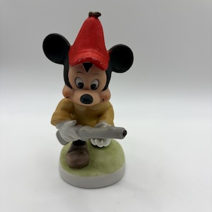 May include: A ceramic figurine of Mickey Mouse wearing a red hat and yellow shirt, holding a gray object. The figurine is standing on a green and white base. The figure has black ears and a black nose.