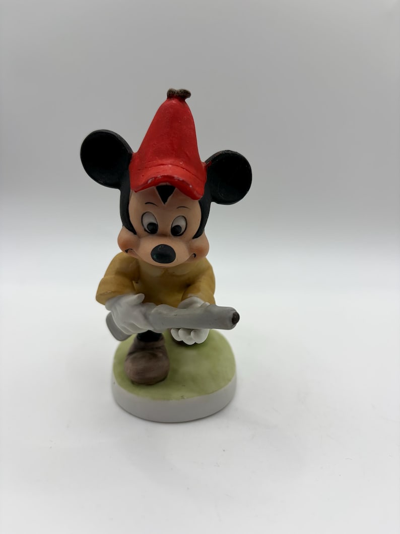 May include: A ceramic figurine of Mickey Mouse wearing a red hat and yellow shirt, holding a gray object. The figurine is standing on a green and white base. The figure has black ears and a black nose.
