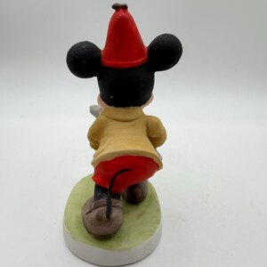 May include: A ceramic figurine of Mickey Mouse from the back, wearing a red hat, yellow coat, and red pants. The figure stands on a green oval base with a white border. The figurine is a collectible item.