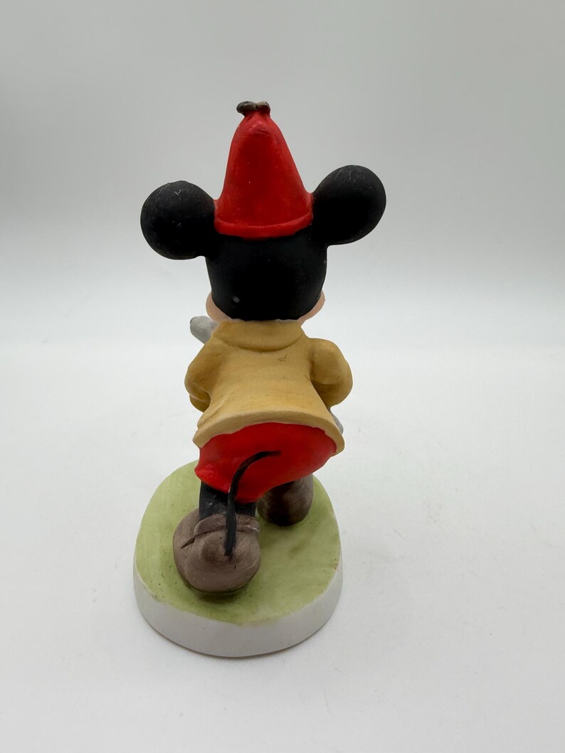 May include: A ceramic figurine of Mickey Mouse from the back, wearing a red hat, yellow coat, and red pants. The figure stands on a green oval base with a white border. The figurine is a collectible item.