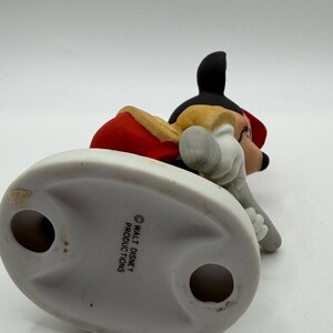 May include: A ceramic figurine of Mickey Mouse in a red jacket, black shorts, and yellow shoes. The figurine is lying down, with its head and arms visible. The base is white and oval-shaped, with the text "WALT DISNEY PRODUCTIONS".