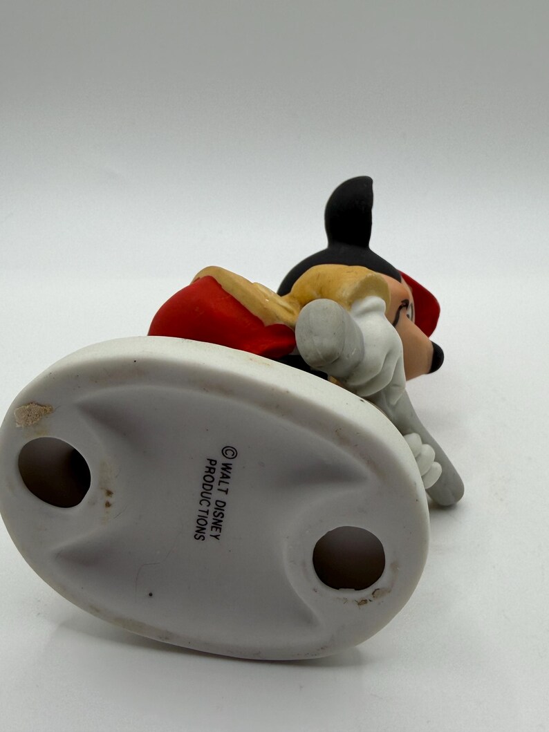 May include: A ceramic figurine of Mickey Mouse in a red jacket, black shorts, and yellow shoes. The figurine is lying down, with its head and arms visible. The base is white and oval-shaped, with the text "WALT DISNEY PRODUCTIONS".