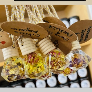 May include: Car diffusers with wooden tops and clear, geometric-shaped glass bottles filled with yellow liquid, dried flowers, and gold flakes. Each diffuser has a small tag with a scent name. The text "Car diffusers" is in a black rectangle.