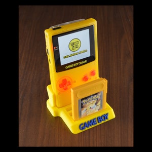 Puede incluir: Una consola Game Boy Color de color amarillo brillante con un soporte a juego, que sostiene un cartucho de juego de Pokémon amarillo. La pantalla muestra el texto "SOLOHEN MODS" y "GAME BOY COLOR". El soporte tiene las palabras "GAME BOY" en azul.