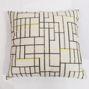 Puede incluir: Una almohada blanca con un patrón geométrico en negro y amarillo. El patrón es una serie de cuadrados y rectángulos superpuestos.