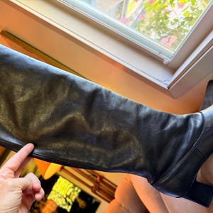 May include: A black knee-high boot crafted from smooth leather, featuring a structured shaft, a rounded toe, and a sturdy block heel. The boot has a clean, minimalist design with visible stitching along the foot and a vertical seam up the side.