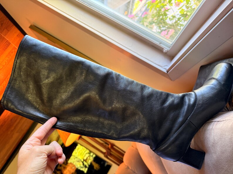 May include: A black knee-high boot crafted from smooth leather, featuring a structured shaft, a rounded toe, and a sturdy block heel. The boot has a clean, minimalist design with visible stitching along the foot and a vertical seam up the side.