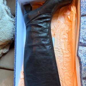 May include: A black leather knee-high boot with a square toe and a block heel, resting inside a rectangular cardboard box lined with light orange tissue paper. The boot features a smooth leather upper with subtle creasing along the shaft.