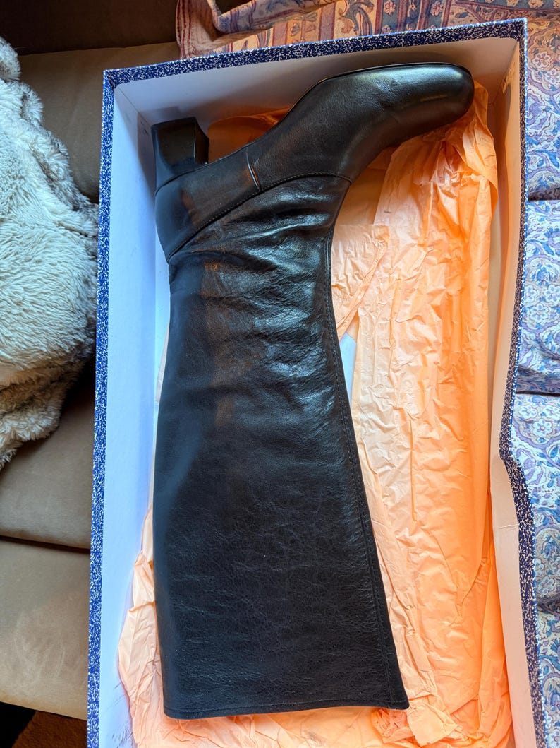 May include: A black leather knee-high boot with a square toe and a block heel, resting inside a rectangular cardboard box lined with light orange tissue paper. The boot features a smooth leather upper with subtle creasing along the shaft.