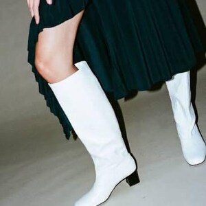 May include: A pair of knee-high white leather boots with a square toe and a low, block-shaped black heel. The boots are worn with a dark green skirt that features a ruffled hemline and a high side slit, revealing the leg above the boot top.