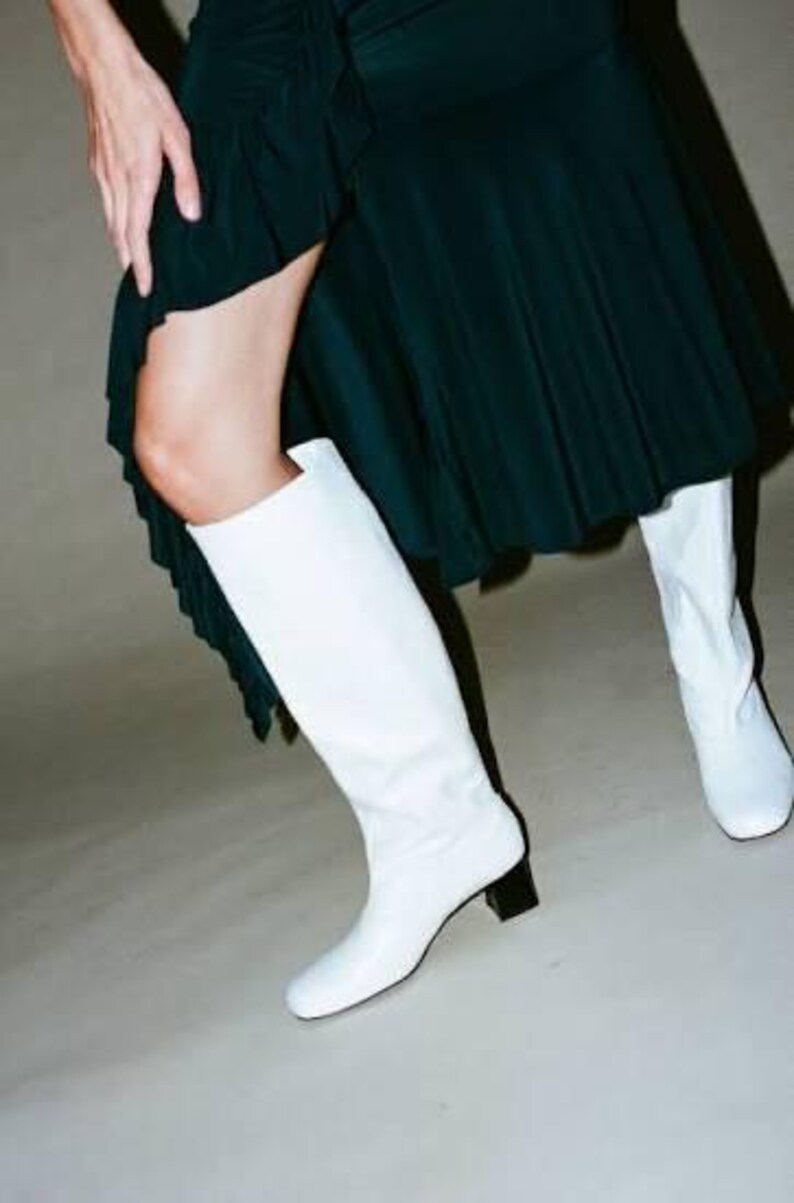 May include: A pair of knee-high white leather boots with a square toe and a low, block-shaped black heel. The boots are worn with a dark green skirt that features a ruffled hemline and a high side slit, revealing the leg above the boot top.