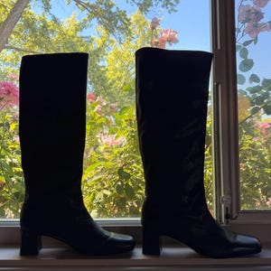 May include: A pair of knee-high black boots with a smooth finish, featuring a rounded toe and a block heel. The boots have a structured shaft that reaches below the knee, with a clean silhouette and no visible hardware or decorative elements.