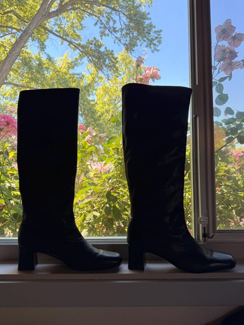 May include: A pair of knee-high black boots with a smooth finish, featuring a rounded toe and a block heel. The boots have a structured shaft that reaches below the knee, with a clean silhouette and no visible hardware or decorative elements.