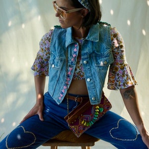 Puede incluir: Una mujer lleva un chaleco vaquero con bordados rosas, una blusa de estampado floral y unos vaqueros azules con adornos en forma de coraz&oacute;n. Est&aacute; sentada en un taburete de madera y lleva un bolso de cuentas de colores.