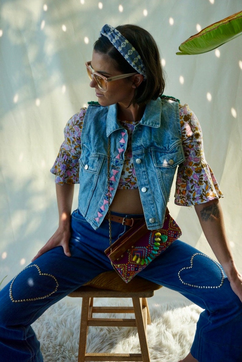 Puede incluir: Una mujer lleva un chaleco vaquero con bordados rosas, una blusa de estampado floral y unos vaqueros azules con adornos en forma de coraz&oacute;n. Est&aacute; sentada en un taburete de madera y lleva un bolso de cuentas de colores.