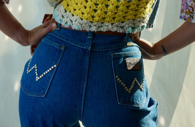 Puede incluir: Un par de pantalones vaqueros azules con un bolsillo trasero con tachuelas y una etiqueta de Wrangler. Los vaqueros tienen cintura alta y pierna ancha.