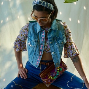 Puede incluir: Una mujer lleva un chaleco vaquero con ribetes rosas y azules, una blusa de estampado floral y unos vaqueros azules con adornos en forma de coraz&oacute;n. Lleva un peque&ntilde;o bolso bordado de colores.