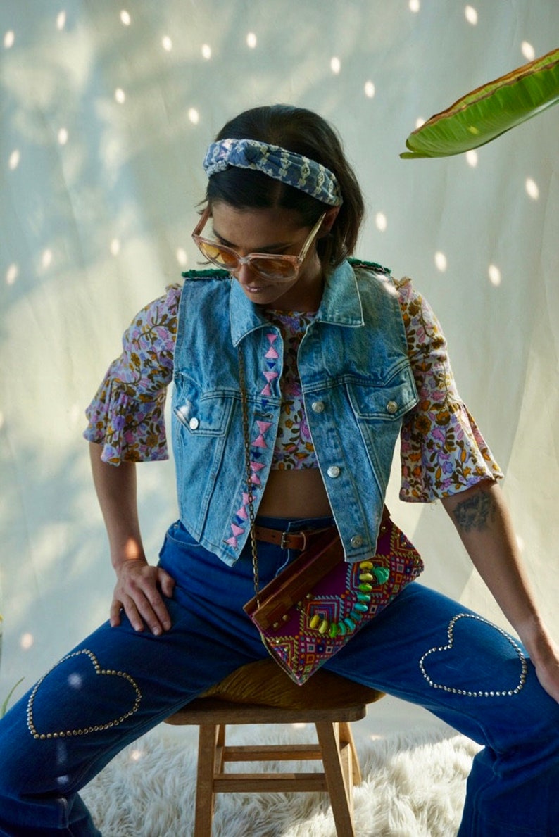 Puede incluir: Una mujer lleva un chaleco vaquero con ribetes rosas y azules, una blusa de estampado floral y unos vaqueros azules con adornos en forma de coraz&oacute;n. Lleva un peque&ntilde;o bolso bordado de colores.