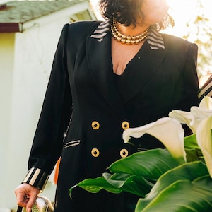 May include: Black double-breasted blazer with gold buttons and striped accents. The blazer is paired with a black top and skirt. Gold heels are held in the hand. A gold necklace is worn.