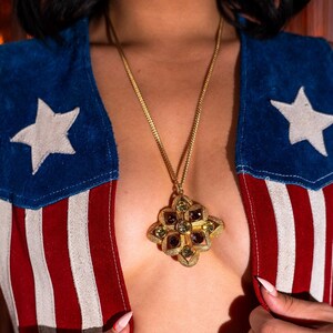 May include: A gold chain necklace with a large, ornate pendant featuring a geometric design with green and purple gemstones. The necklace is worn by a person wearing a red, white, and blue patterned jacket.