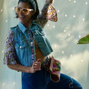 Puede incluir: Una mujer con un chaleco vaquero con bordados rosas y azules, una blusa floral, pantalones vaqueros azules y un bolso de cuentas de colores. Lleva gafas de sol y una diadema.