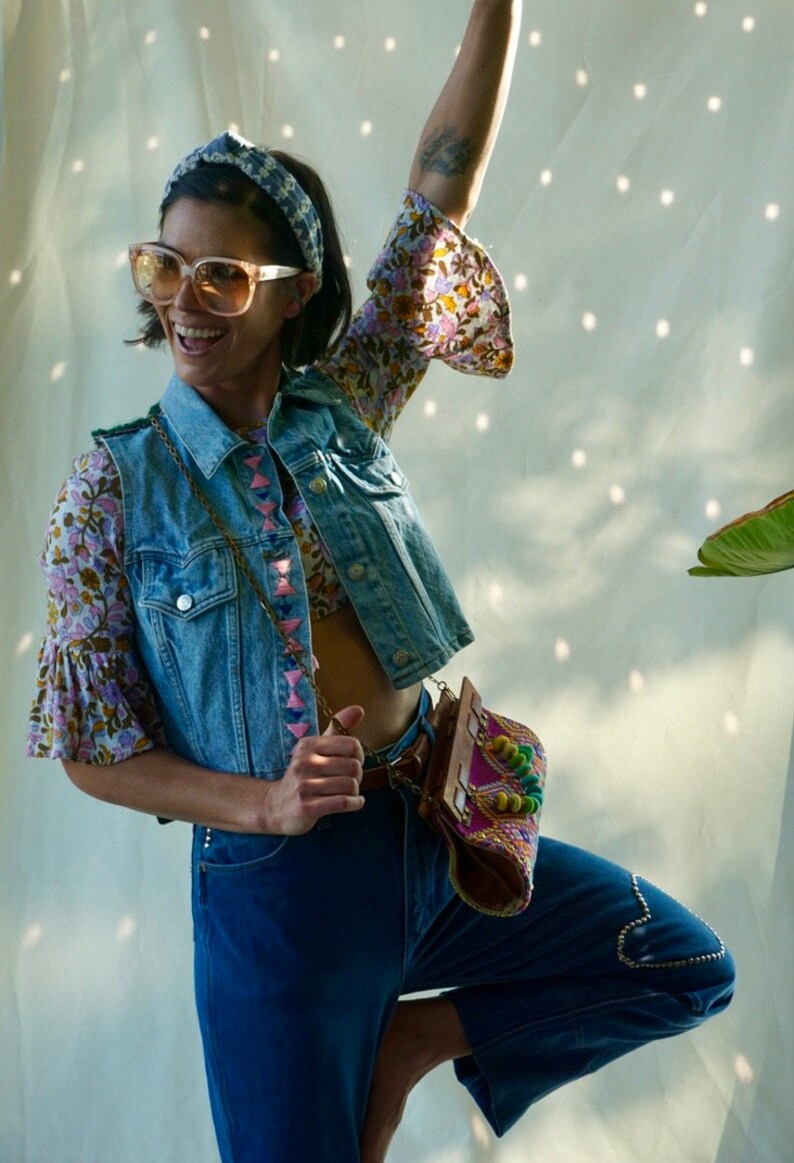 Puede incluir: Una mujer con un chaleco vaquero con bordados rosas y azules, una blusa floral, pantalones vaqueros azules y un bolso de cuentas de colores. Lleva gafas de sol y una diadema.